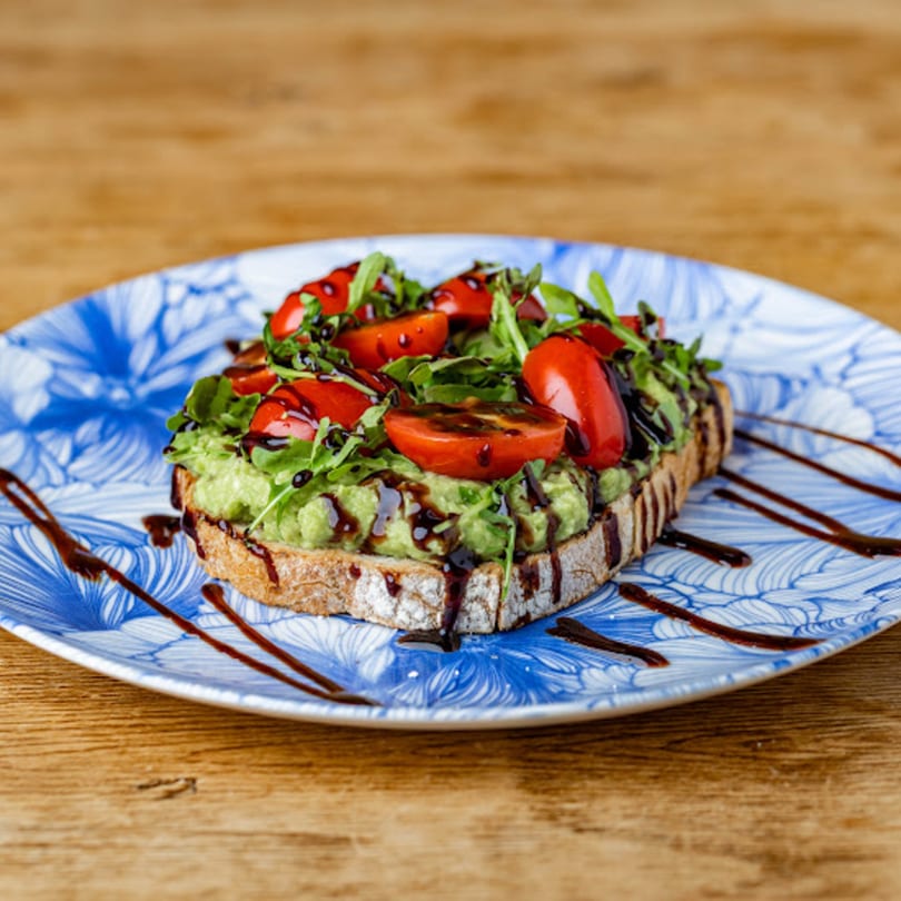 Avocado toast with cherry tomatoes, arugula, and balsamic glaze on a blue and white plate.
